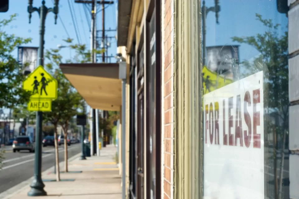 Close-up of For Lease sign in window of shop storefront with city sidewalk and vintage street light in background.